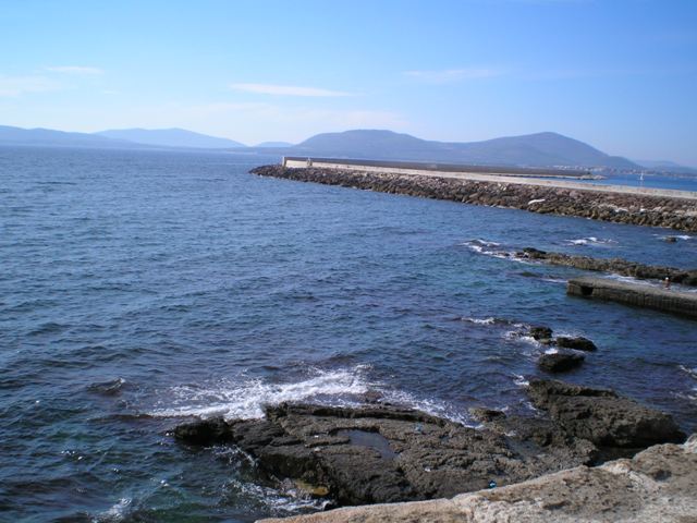 Dalla costa di Alghero si vede il molo e qualche scoglio bagnato dal mare che spumeggia infrangendosi sulle rocce. In lontananza una collina si fa timidamente vedere.
